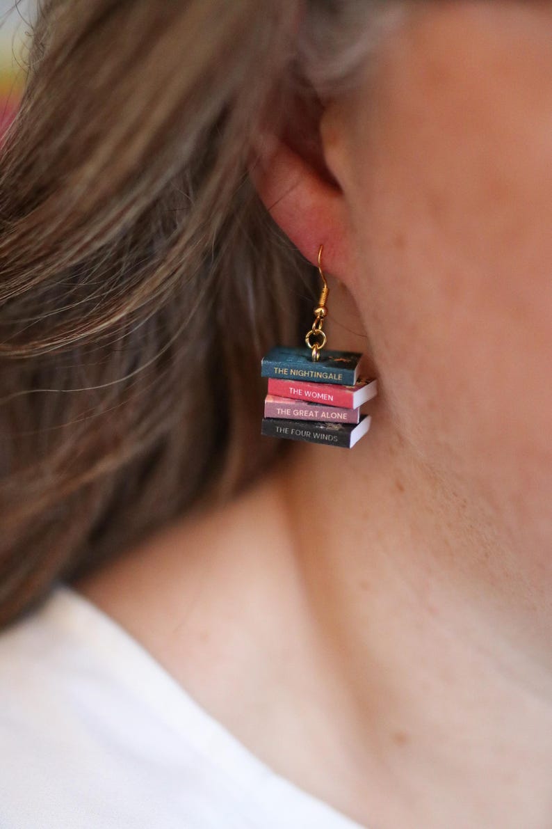 Puede incluir: Pendientes colgantes dorados con mini pilas de libros. Cada libro tiene un t&iacute;tulo diferente: "The Nightingale", "The Women", "The Great Alone" y "The Four Winds". Un accesorio &uacute;nico para los amantes de los libros.