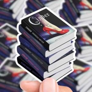 Könnte beinhalten: Ein Stapel Aufkleber mit den Buchcovern aus Marissa Meyers Lunar Chronicles-Reihe. Der oberste Aufkleber zeigt das Buch "Cinder" mit einem roten Schuh. Die anderen Aufkleber zeigen die Titel "Scarlet", "Cress", "Fairest", "Winter" und "Stars Above".