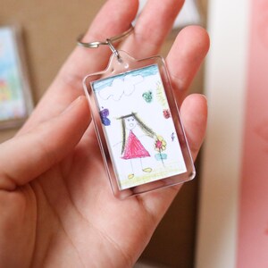 Peut inclure: Un porte-cl&eacute;s en plastique transparent avec un dessin d'une fille en robe rouge et une fleur. Le dessin est entour&eacute; d'une bordure blanche.