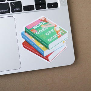Puede incluir: Una pila de tres pegatinas de libros sobre un port&aacute;til. El libro superior es verde con el t&iacute;tulo "Nora Goes Off Script". El libro del medio es azul y el libro inferior es rojo.