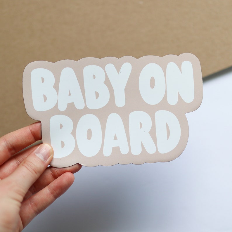 May include: A light pink car sign with white text that reads "BABY ON BOARD" in a bubble font.