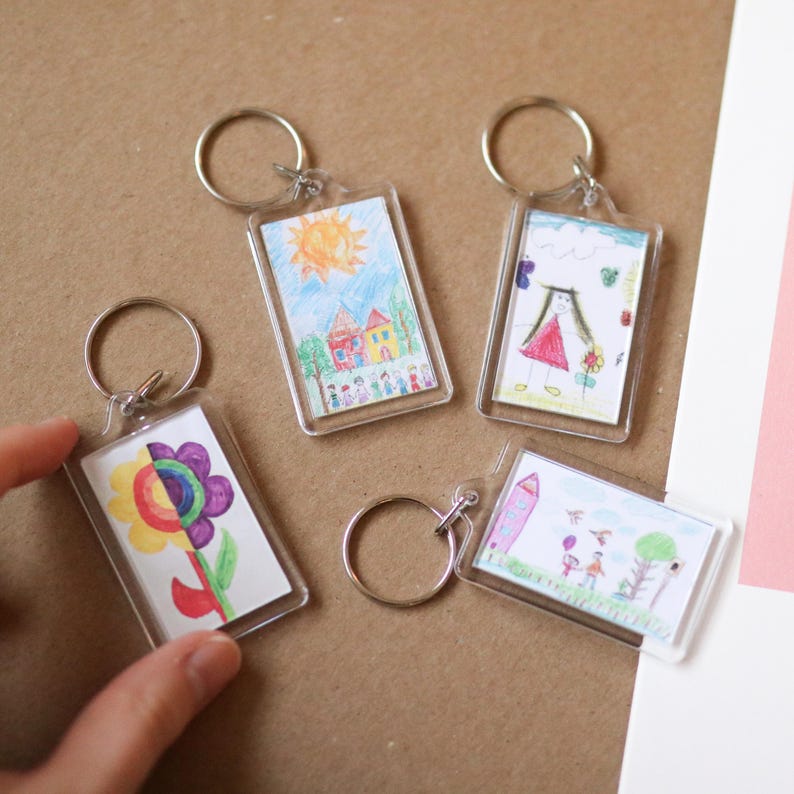 Peut inclure: Quatre porte-cl&eacute;s en plastique transparent avec des dessins color&eacute;s &agrave; l'int&eacute;rieur. Les dessins comprennent une maison avec un soleil, une fille en robe rouge, une fleur et une fille et un gar&ccedil;on dans un parc.
