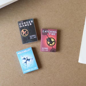May include: A set of miniature books, including "The Hunger Games," "Catching Fire," and "Mockingjay" by Suzanne Collins. Each book displays a distinct cover design with a bird emblem and title text. The books are in black, red, and blue.