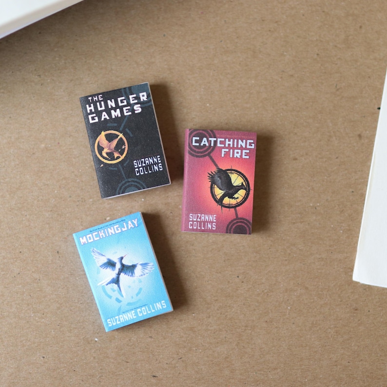 May include: A set of miniature books, including "The Hunger Games," "Catching Fire," and "Mockingjay" by Suzanne Collins. Each book displays a distinct cover design with a bird emblem and title text. The books are in black, red, and blue.