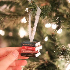 以下が含まれることがあります： クリスマスツリーのオーナメント。ミニチュアの本が積み重ねられており、「The Nightingale」、「The Women」、「The Great Alone」、「The Four Winds」などのタイトルが見える。白いリボンで吊り下げられている。