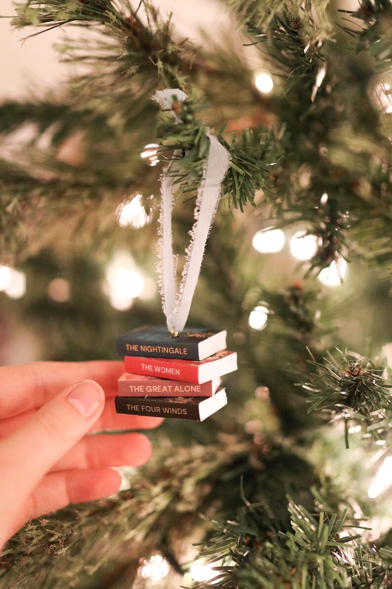 以下が含まれることがあります： クリスマスツリーのオーナメント。ミニチュアの本が積み重ねられており、「The Nightingale」、「The Women」、「The Great Alone」、「The Four Winds」などのタイトルが見える。白いリボンで吊り下げられている。