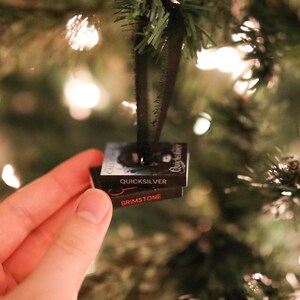 May include: A miniature Christmas tree ornament shaped like two stacked books. The top book reads "Quicksilver" and the bottom book reads "Brimstone". The ornament is black with red and white text, and is hanging from a black ribbon.