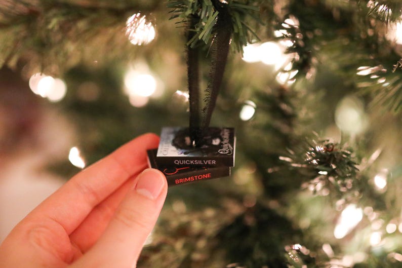May include: A miniature Christmas tree ornament shaped like two stacked books. The top book reads "Quicksilver" and the bottom book reads "Brimstone". The ornament is black with red and white text, and is hanging from a black ribbon.