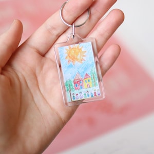 Peut inclure: Un porte-cl&eacute;s en plastique transparent avec un dessin d'un soleil, des maisons et des personnes &agrave; l'int&eacute;rieur. Le dessin est color&eacute; et semble avoir &eacute;t&eacute; fait par un enfant.