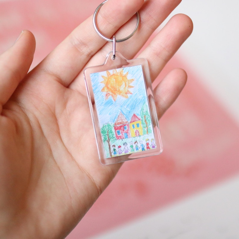 Peut inclure: Un porte-cl&eacute;s en plastique transparent avec un dessin d'un soleil, des maisons et des personnes &agrave; l'int&eacute;rieur. Le dessin est color&eacute; et semble avoir &eacute;t&eacute; fait par un enfant.