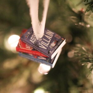 K&ouml;nnte beinhalten: Ein Stapel Miniatur-Buchornamente, die an einem wei&szlig;en Band h&auml;ngen. Das oberste Buch ist lila und tr&auml;gt den Titel "ASSASSIN TO THE VILLAIN". Die anderen B&uuml;cher sind rot und blau. Die Ornamente h&auml;ngen vor einem verschwommenen Weihnachtsbaum.