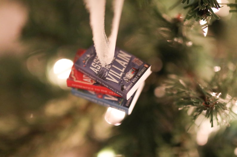 K&ouml;nnte beinhalten: Ein Stapel Miniatur-Buchornamente, die an einem wei&szlig;en Band h&auml;ngen. Das oberste Buch ist lila und tr&auml;gt den Titel "ASSASSIN TO THE VILLAIN". Die anderen B&uuml;cher sind rot und blau. Die Ornamente h&auml;ngen vor einem verschwommenen Weihnachtsbaum.