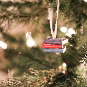 K&ouml;nnte beinhalten: Weihnachtsbaumschmuck mit drei kleinen B&uuml;chern mit den Titeln "Assistant to the Villain", "Apprentice to the Villain" und "Accomplice to the Villain". Die B&uuml;cher sind an einem wei&szlig;en Band aufgeh&auml;ngt, vor einem Weihnachtsbaum.