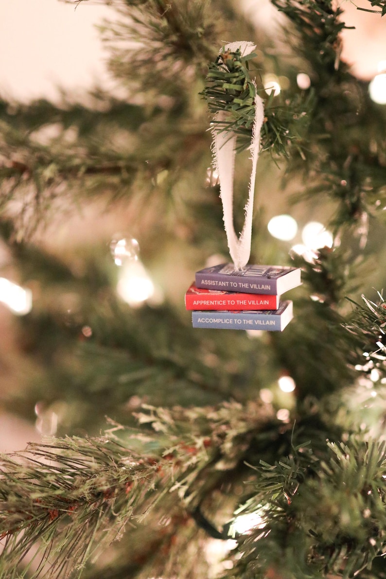 K&ouml;nnte beinhalten: Weihnachtsbaumschmuck mit drei kleinen B&uuml;chern mit den Titeln "Assistant to the Villain", "Apprentice to the Villain" und "Accomplice to the Villain". Die B&uuml;cher sind an einem wei&szlig;en Band aufgeh&auml;ngt, vor einem Weihnachtsbaum.
