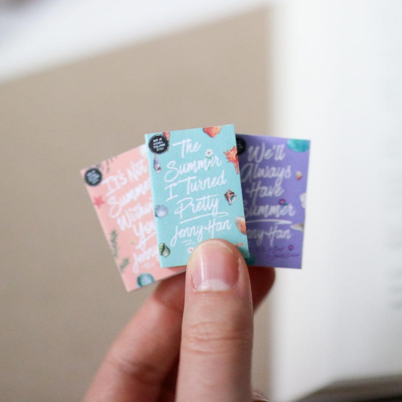 May include: Three miniature book covers with titles including "It's Not Summer Without You", "The Summer I Turned Pretty", and "We'll Always Have Summer" by Jenny Han. The covers are pastel colors with decorative elements.