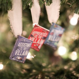 May include: Three miniature book ornaments hang from a Christmas tree. The books have titles like "Assistant to the Villain" and "Apprentice to the Villain." Each book is attached to a white ribbon.