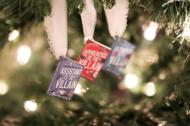 May include: Three miniature book ornaments hang from a Christmas tree. The books have titles like "Assistant to the Villain" and "Apprentice to the Villain." Each book is attached to a white ribbon.