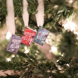 May include: Three miniature book ornaments hang from a Christmas tree. The books are titled "Assistant to the Villain," "Apprentice to the Villain," and "Accomplice to the Villain." The ornaments are blue, red, and purple.