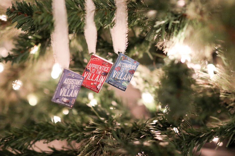 May include: Three miniature book ornaments hang from a Christmas tree. The books are titled "Assistant to the Villain," "Apprentice to the Villain," and "Accomplice to the Villain." The ornaments are blue, red, and purple.