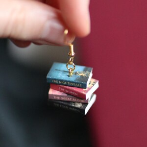 Puede incluir: Un pendiente dorado con una pila de libros en miniatura. Los libros tienen t&iacute;tulos como "The Nightingale", "The Women", "The Great Alone" y "The Four Winds". Los libros son de color azul, rojo y negro.