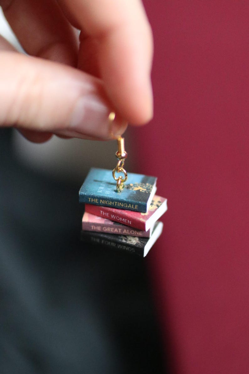Puede incluir: Un pendiente dorado con una pila de libros en miniatura. Los libros tienen t&iacute;tulos como "The Nightingale", "The Women", "The Great Alone" y "The Four Winds". Los libros son de color azul, rojo y negro.