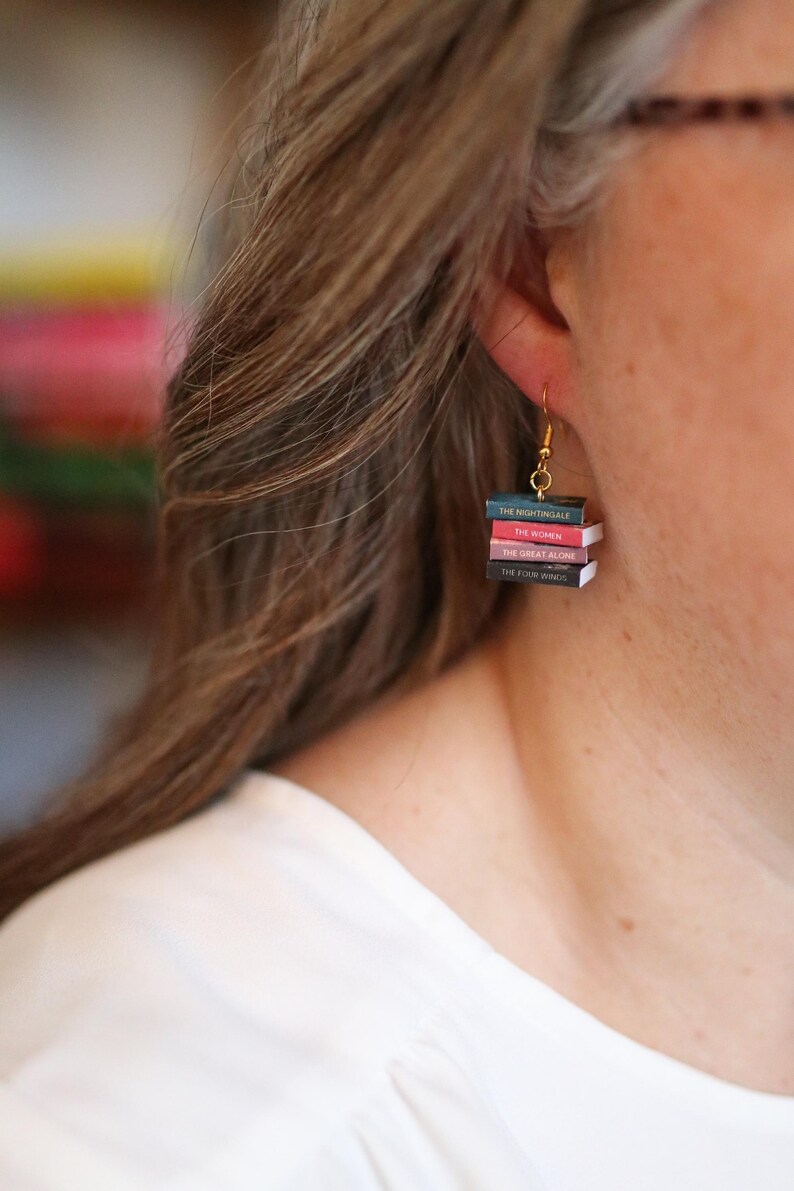 Puede incluir: Pendientes colgantes dorados con forma de pilas de libros en miniatura. Cada libro tiene un lomo de diferente color y t&iacute;tulo: "The Nightingale", "The Women", "The Great Alone" y "The Four Winds". Un accesorio original para amantes de los libros.