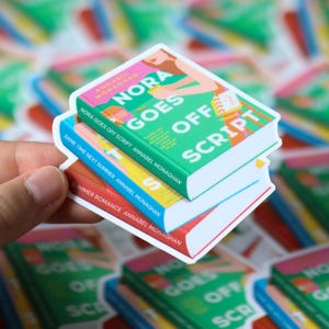 Puede incluir: Tres pegatinas con forma de libro apiladas. La pegatina superior es verde con el t&iacute;tulo "Nora Goes Off Script". La pegatina del medio es azul y amarilla, y la inferior es roja. El nombre del autor tambi&eacute;n es visible.