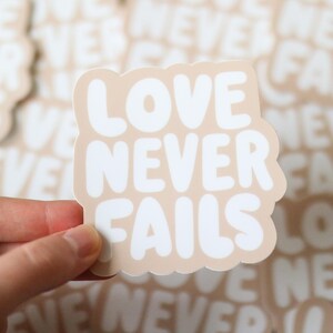Peut inclure: Un autocollant beige avec les mots "LOVE NEVER FAILS" en blanc, en gras et en lettres arrondies. L'autocollant a une forme douce en forme de nuage. D'autres autocollants avec le même design sont visibles en arrière-plan.