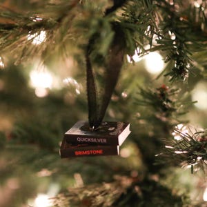 May include: A miniature Christmas ornament shaped like two stacked books. The top book is titled "Quicksilver" and the bottom book is titled "Brimstone". The ornament is suspended from a black ribbon and set against a backdrop of a Christmas tree.