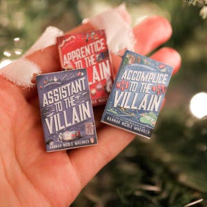 May include: Three miniature books are held in a hand, with titles including "Assistant to the Villain" and "Accomplice to the Villain". The books have colourful covers with text and illustrations. The background is blurred with Christmas tree branches.