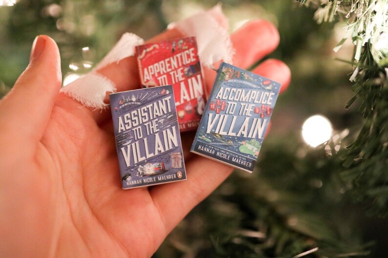 May include: Three miniature books are held in a hand, with titles including "Assistant to the Villain" and "Accomplice to the Villain". The books have colourful covers with text and illustrations. The background is blurred with Christmas tree branches.