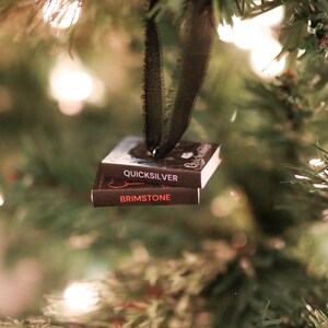 May include: A Christmas tree ornament shaped like two miniature books, "Quicksilver" and "Brimstone", suspended by a black ribbon. The books are dark with white text and red accents, hanging against a backdrop of green pine needles and soft, out-of-focus lights.