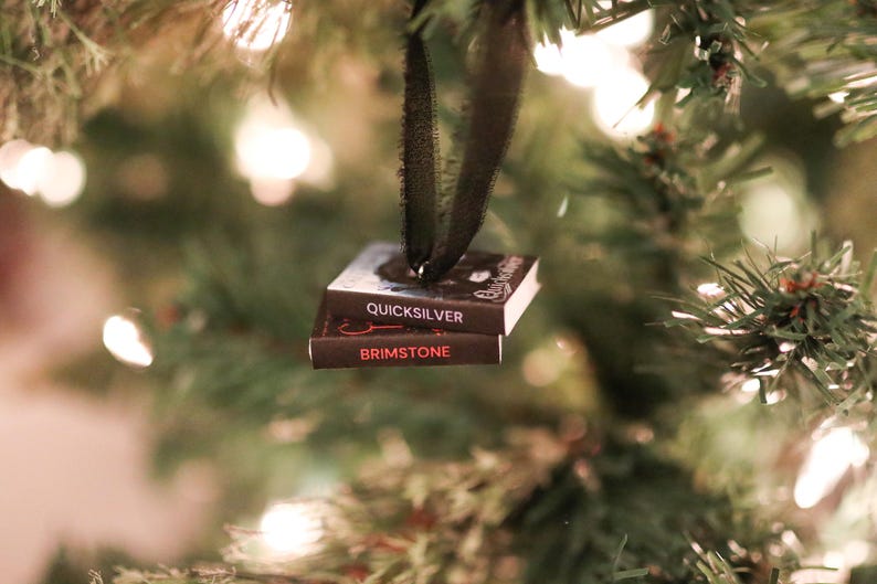 May include: A Christmas tree ornament shaped like two miniature books, "Quicksilver" and "Brimstone", suspended by a black ribbon. The books are dark with white text and red accents, hanging against a backdrop of green pine needles and soft, out-of-focus lights.