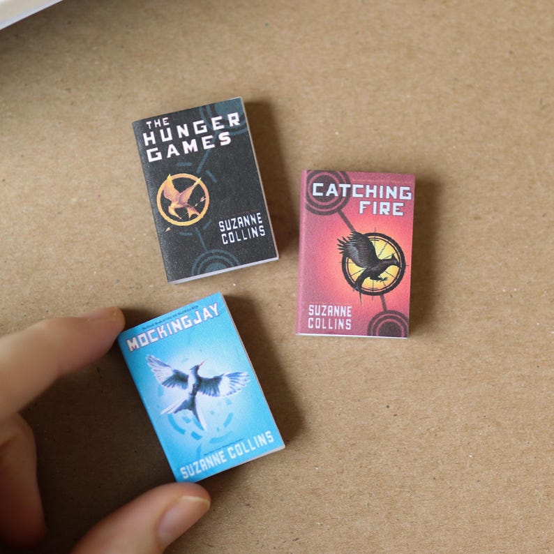May include: A set of miniature books, including "The Hunger Games," "Catching Fire," and "Mockingjay" by Suzanne Collins. Each book has a distinct cover design with a mockingjay and flame. The author's name and book titles are visible.
