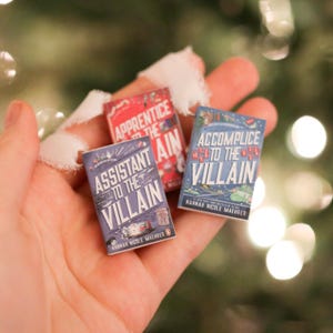 May include: Three miniature books with colourful covers are held in a hand. The books are titled "Assistant to the Villain", "Apprentice to the Villain", and "Accomplice to the Villain". The author's name, Hannah Nicole Maehrer, is printed on the bottom of two of the books.