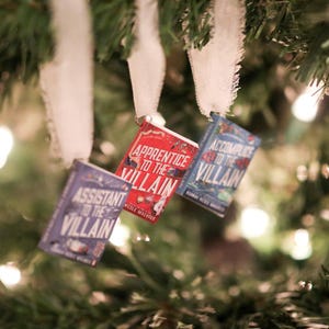 May include: Three miniature book ornaments hang from a Christmas tree. The books are titled "Assistant to the Villain", "Apprentice to the Villain", and "Accomplice to the Villain". The books are in shades of red, blue, and purple.
