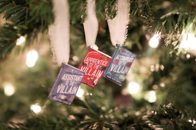 May include: Three miniature book ornaments hang from a Christmas tree. The books are titled "Assistant to the Villain", "Apprentice to the Villain", and "Accomplice to the Villain". The books are in shades of red, blue, and purple.
