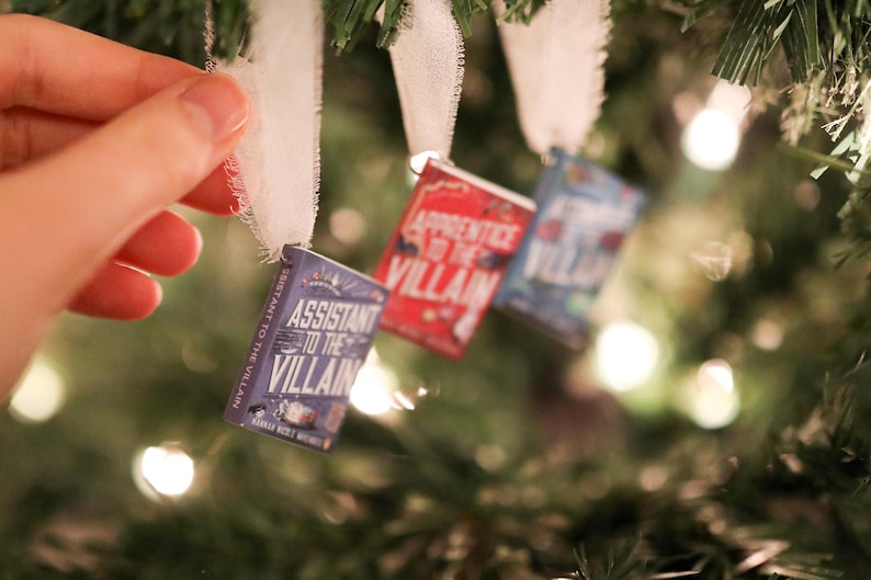 May include: Miniature book ornaments hanging on a Christmas tree. The ornaments are small, rectangular, and feature book covers with titles like "Assistant to the Villain" and "Apprentice to the Villain". Each book is attached to a white ribbon.