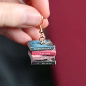 Puede incluir: Un pendiente dorado con una pila de libros en miniatura. Los libros se titulan "The Nightingale", "The Women", "The Great Alone" y "The Four Winds". El pendiente se muestra sobre un fondo rojo oscuro.