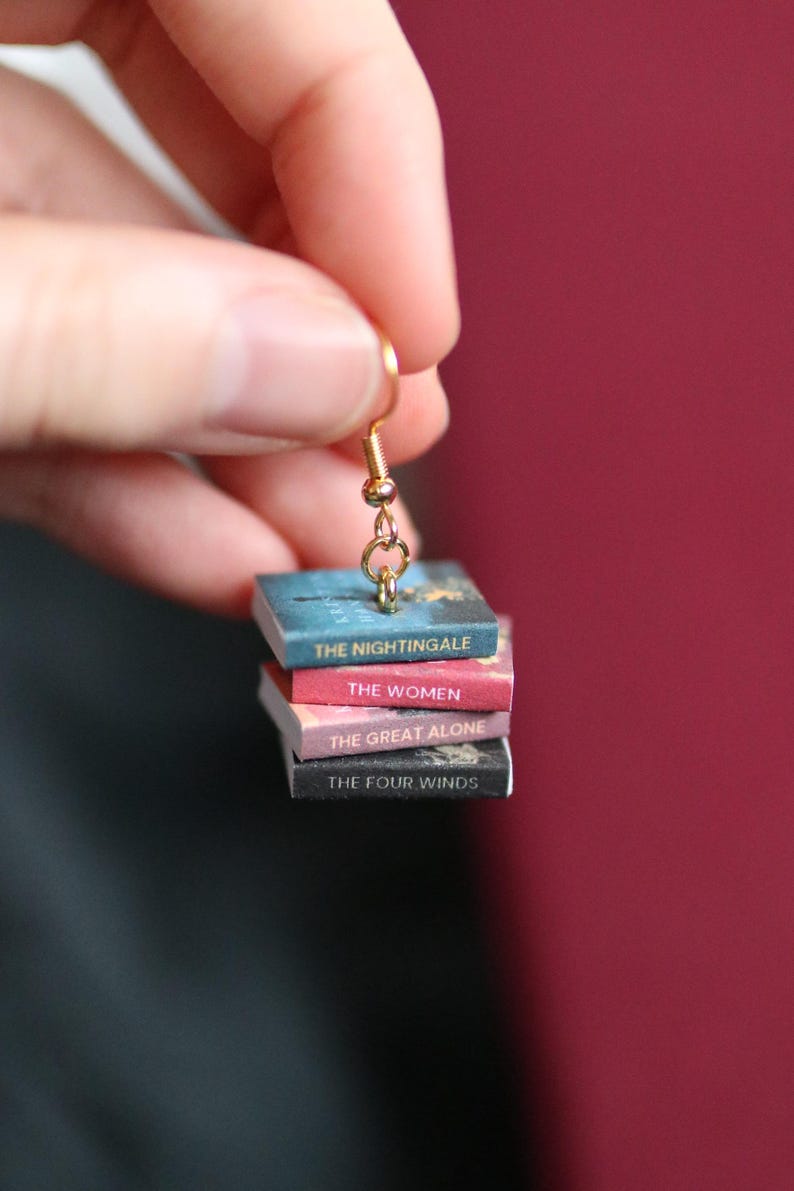 Puede incluir: Un pendiente dorado con una pila de libros en miniatura. Los libros se titulan "The Nightingale", "The Women", "The Great Alone" y "The Four Winds". El pendiente se muestra sobre un fondo rojo oscuro.