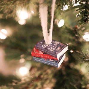 K&ouml;nnte beinhalten: Ein Weihnachtsbaumschmuck mit drei Miniaturb&uuml;chern. Die B&uuml;cher tragen die Titel "Assistant to the Villain", "Apprentice to the Villain" und "Accomplice to the Villain". Die B&uuml;cher sind blau und rot und werden von einem wei&szlig;en Band gehalten.