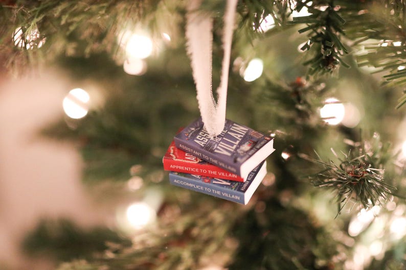 K&ouml;nnte beinhalten: Ein Weihnachtsbaumschmuck mit drei Miniaturb&uuml;chern. Die B&uuml;cher tragen die Titel "Assistant to the Villain", "Apprentice to the Villain" und "Accomplice to the Villain". Die B&uuml;cher sind blau und rot und werden von einem wei&szlig;en Band gehalten.