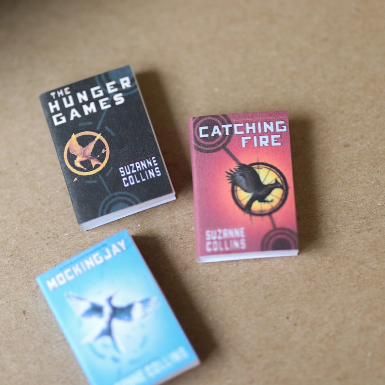 May include: Three miniature books from The Hunger Games series. The books are titled "The Hunger Games", "Catching Fire", and "Mockingjay". Each book features a different cover design with the author's name, Suzanne Collins.