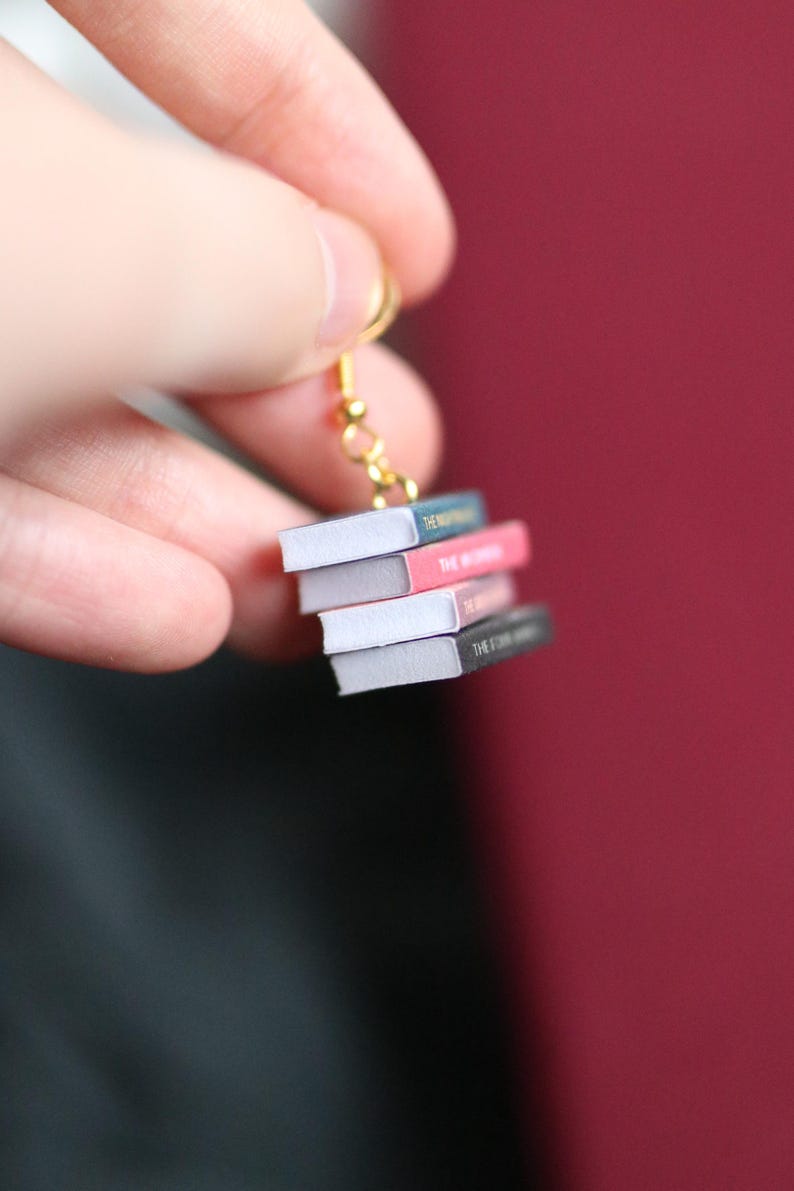 Puede incluir: Pendiente dorado con una pila de libros en miniatura. Los libros tienen p&aacute;ginas blancas y lomos coloridos en tonos azules, rosas y grises. El texto "THE" y "THE WORDS" es visible en los lomos. El pendiente est&aacute; sobre un fondo burdeos.