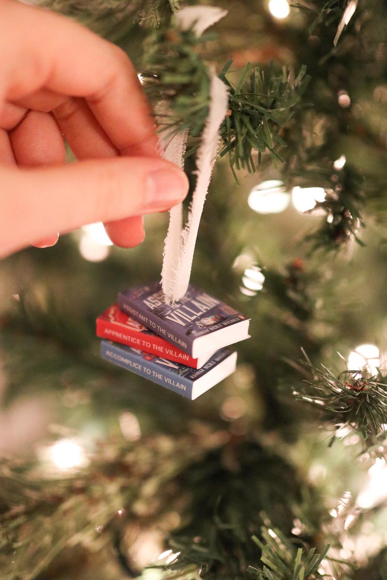 K&ouml;nnte beinhalten: Weihnachtsbaumschmuck mit drei Miniaturb&uuml;chern. Die B&uuml;cher sind gestapelt und tragen Titel wie "Assistant to the Villain", "Apprentice to the Villain" und "Accomplice to the Villain". Die B&uuml;cher sind rot, blau und lila.