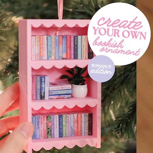 Könnte beinhalten: Ein rosafarbenes Miniatur-Bücherregal-Ornament mit drei gewellten Regalböden, gefüllt mit winzigen bunten Buchrücken und einer kleinen Topfpflanze. Das Ornament hat ein rosa Band zum Aufhängen und den Text create YOUR OWN bookish ornament ROMANCE edition.