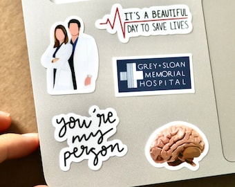 May include: A laptop computer with several stickers on it. The stickers include a heart with a heartbeat line, the text "It's a beautiful day to save lives", a blue and white sticker with the text "Grey Sloan Memorial Hospital", a sticker with the text "you're my person", and a sticker of a human brain.