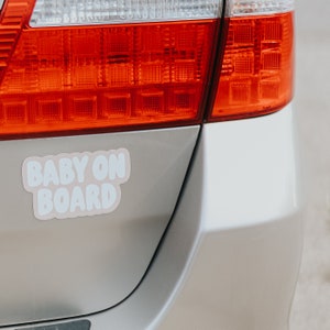 May include: A silver car with a pink and white "Baby On Board" sticker on the back bumper. The car has a red taillight.