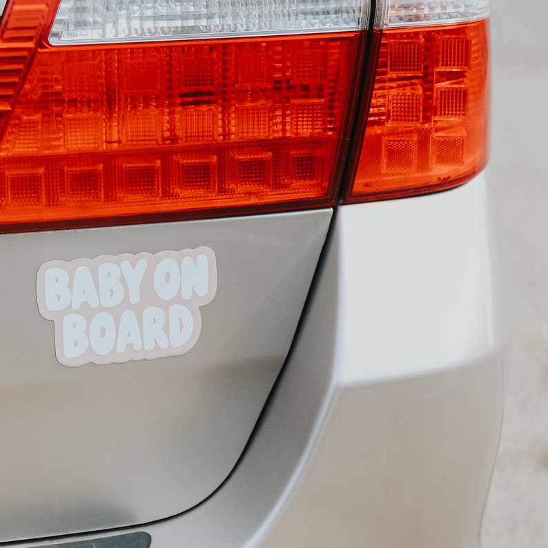May include: A silver car with a pink and white "Baby On Board" sticker on the back bumper. The car has a red taillight.