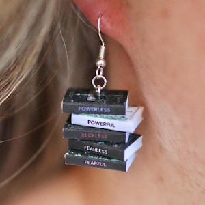 May include: Silver-tone dangle earrings featuring a stack of miniature books. Each book has a different title: "Powerless," "Powerful," "Reckless," "Fearless," and "Fearful." The earrings are designed to resemble a small stack of books.